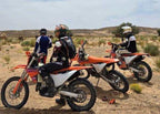 Three orange and white off-road dirt bikes with riders in protective gear ready for desert terrain in Moto KTM Rides in Merzouga.