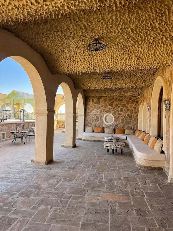 Outdoor seating area at Nasser Palace Suites & Spa featuring beige archways, textured ceiling, stone wall, and long cushioned bench with orange pillows