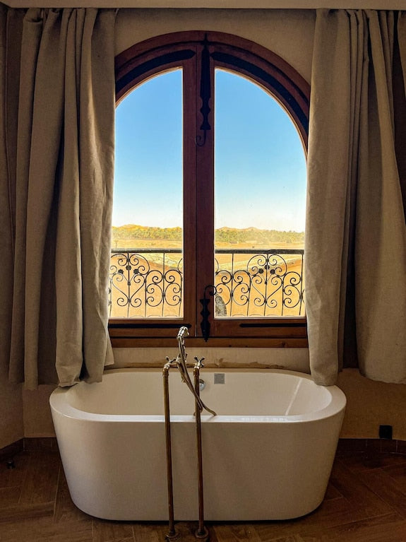 Freestanding white bathtub with chrome faucets framed by beige curtains and a large arched window overlooking a desert landscape at Nasser Palace Suites & Spa