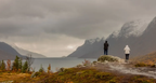 Norway Tromso fjord landscape with two hikers on rocky terrain, snow-capped mountains and autumn vegetation