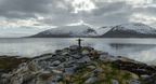 Norway Tromso fjord with rocky shoreline, calm water, snow-covered mountains, and a person standing on rocks