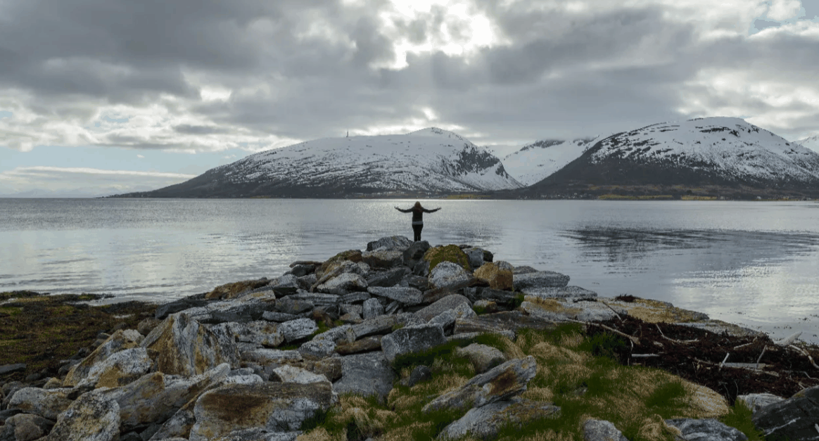 Norway Tromso fjord with rocky shoreline, calm water, snow-covered mountains, and a person standing on rocks