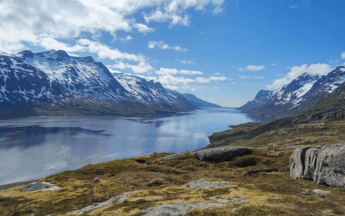Norway Tromso fjord landscape with calm blue water, snow-capped mountains, rocky shoreline, and green tundra