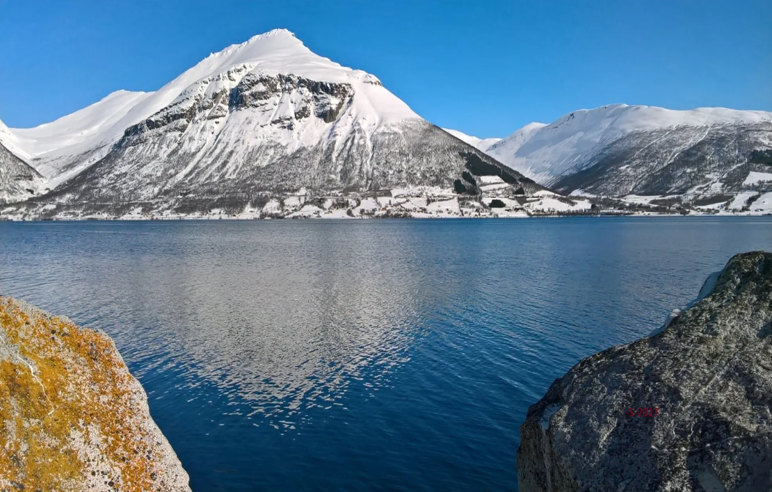 Norway Tromso fjord with snow-covered mountains, blue water, and rocky shoreline in winter