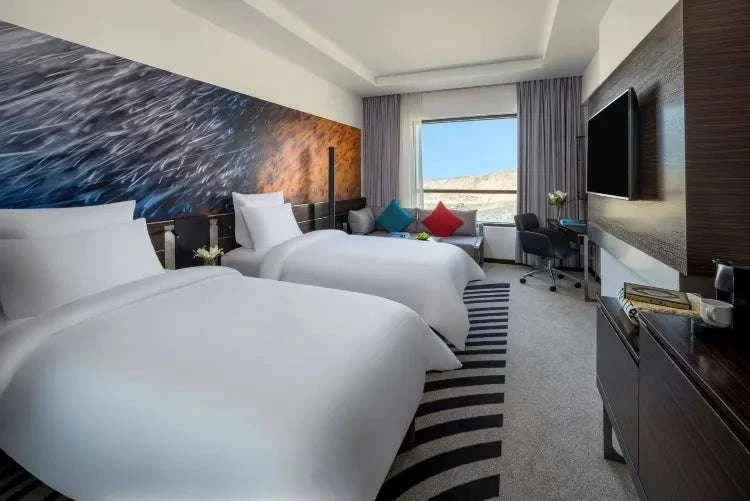 Novotel Makkah Thakher City twin hotel room with white beds, colourful cushions, wall art, TV and mountain view