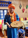 Man in blue traditional Moroccan robe and orange turban holding a round baked Berber pizza on a wooden tray inside a decorated restaurant to Order Pizza.