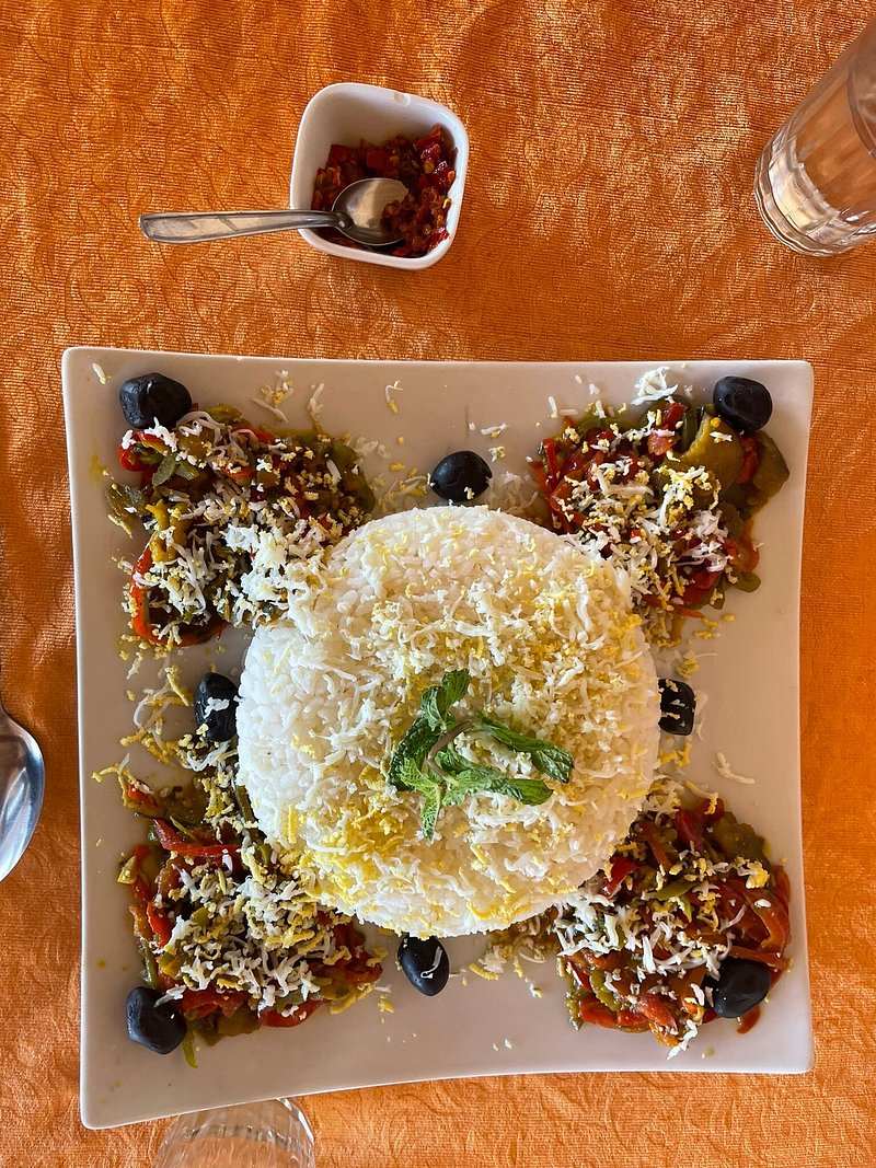 Order Pizza served with a round mound of white rice topped with herbs, surrounded by mixed vegetable salad and black olives on a square plate