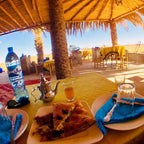 Outdoor dining setup with yellow tablecloth, Moroccan-style teapot, orange juice, and Berber pizza slices under a thatched roof at Restaurant Nora where you can Order Pizza.