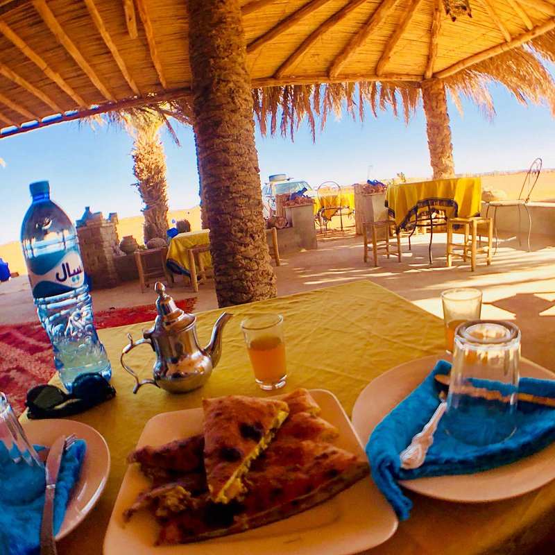 Outdoor dining setup with yellow tablecloth, Moroccan-style teapot, orange juice, and Berber pizza slices under a thatched roof at Restaurant Nora where you can Order Pizza.