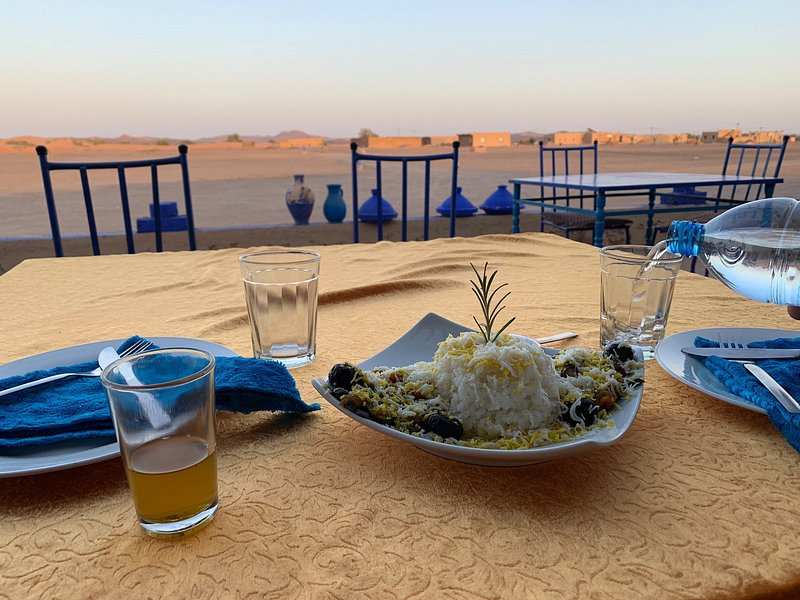 Outdoor dining table with yellow tablecloth, white rice dish garnished with olives and herbs, blue napkins, and clear drinking glasses in desert setting, perfect to Order Pizza in Merzouga.