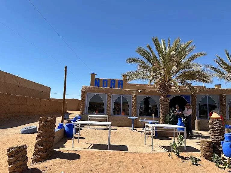 Restaurant building with beige stone walls, blue painted tables and pots, palm tree, and arched windows under a clear blue sky, inviting visitors to Order Pizza.