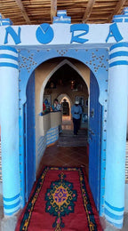 Restaurant entrance painted in blue and white stripes with a decorative archway and a colorful patterned rug on the floor inviting to Order Pizza.