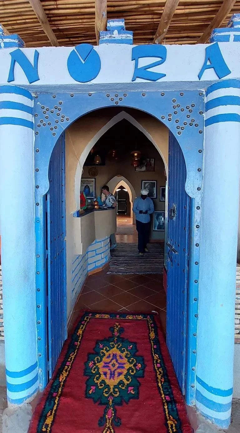 Restaurant entrance painted in blue and white stripes with a decorative archway and a colorful patterned rug on the floor inviting to Order Pizza.