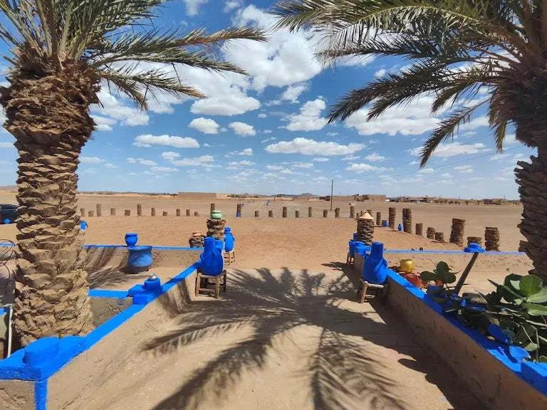 Outdoor desert restaurant setting with palm trees, blue-painted low walls, and clay pots along seating areas, inviting visitors to Order Pizza.