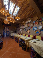 Restaurant dining area with beige tablecloths, patterned cushioned stools, colorful woven baskets on walls, and hanging wicker lamps, inviting to Order Pizza.