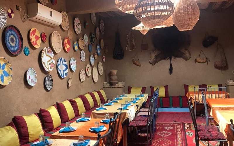 Restaurant interior with long tables covered in orange and beige cloths, colorful cushions, and woven wall baskets in warm earthy tones inviting to Order Pizza.