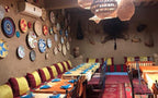 Restaurant interior with long tables covered in orange and beige cloths, colorful cushions, and woven wall baskets in warm earthy tones inviting to Order Pizza.