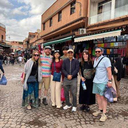 Group of six casually dressed tourists standing on cobblestone street in Marrakech market with red buildings in the background, highlighting Our Representative in Marrakech.