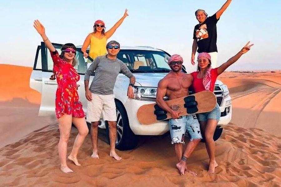 Group of six people posing barefoot on orange sand dunes near a white 4x4 SUV, with one man holding a wooden electric skateboard, wearing casual summer clothing and red scarves, illustrating a Private Guide for Driving experience in the Erg Chebbi desert.