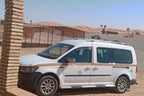 White and black van with tinted windows parked near desert dunes and shaded structure, offering Private Taxi from Erfoud to Merzouga service.