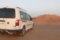 White Volkswagen van parked on desert terrain with reddish sand dunes in the background, ideal for Private Taxi from Erfoud to Merzouga services.