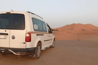 White Volkswagen van parked on desert terrain with reddish sand dunes in the background, ideal for Private Taxi from Erfoud to Merzouga services.