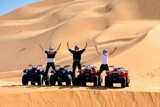 Quad bike ride with three riders on red and blue four-wheelers in beige sandy Merzouga dunes under clear sky
