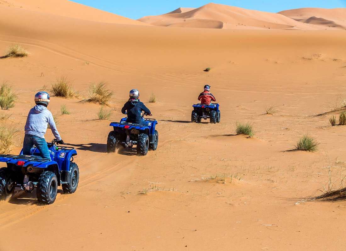 Three people wearing helmets on blue quad bikes riding through sandy orange dunes with sparse green shrubs during a quad bike ride in the desert.