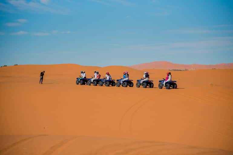 Group of people on blue quad bikes riding across orange sand dunes under a clear blue sky during a quad bike ride in the desert.