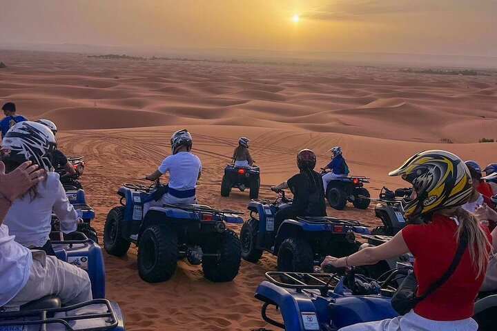 Group of people on blue quad bikes wearing helmets during a sunset quad bike ride across golden sand dunes in the desert.