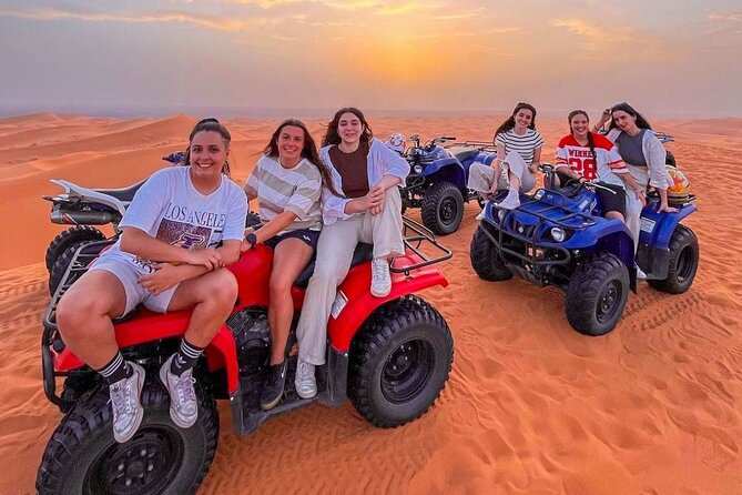 Group of six women enjoying a quad bike ride on red and blue all-terrain vehicles across golden Merzouga sand dunes at sunset.
