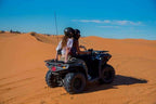 Quad bike ride with two riders wearing helmets on a black ATV in golden sandy desert dunes under clear blue sky.