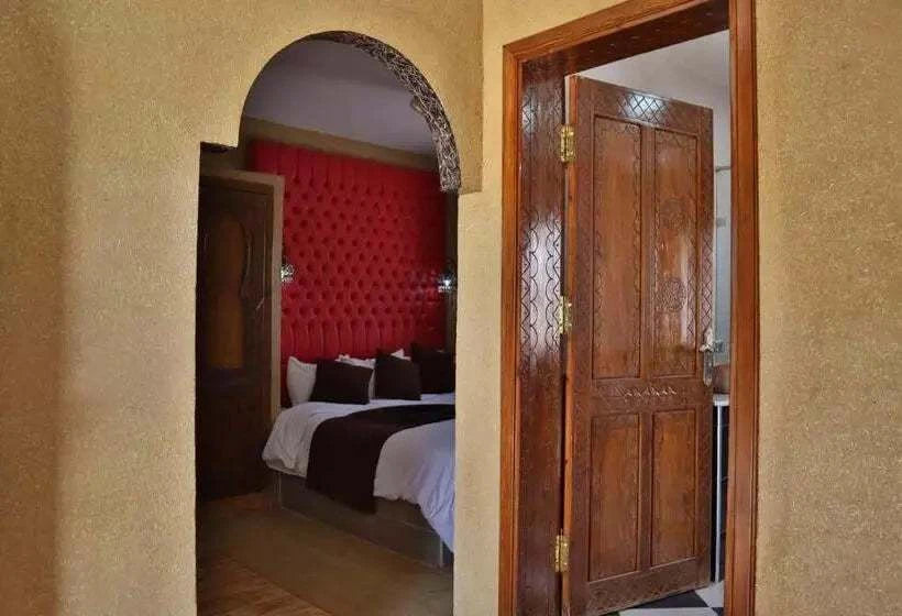 Residence Touristique Merzouga bedroom with beige textured walls, carved wooden door, red tufted headboard, and white bedding with brown accents