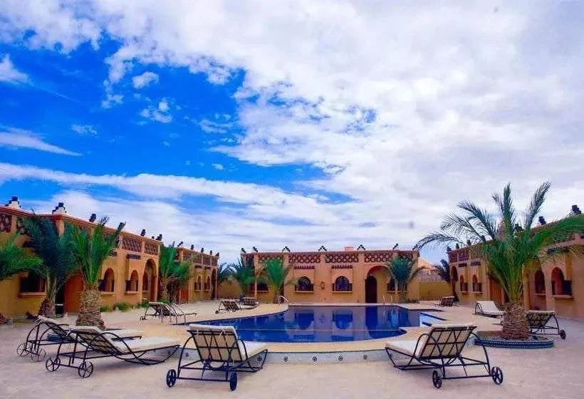 Residence Touristique Merzouga with beige stucco buildings surrounding a central blue swimming pool, black metal lounge chairs, and palm trees under a partly cloudy sky