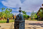 Residence Touristique Merzouga featuring black ornate metal lantern on paved courtyard under a blue sky with surrounding greenery and buildings