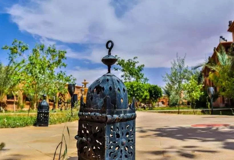 Residence Touristique Merzouga featuring black ornate metal lantern on paved courtyard under a blue sky with surrounding greenery and buildings