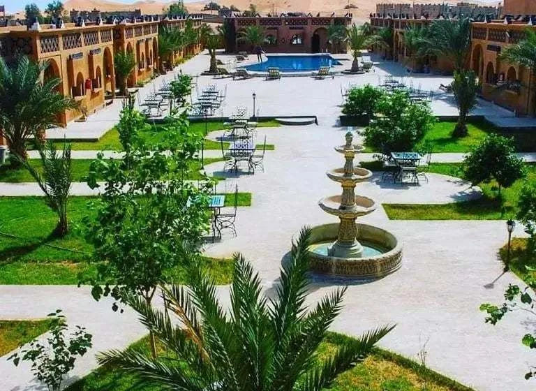 Residence Touristique Merzouga showcasing a spacious courtyard with green lawns, palm trees, outdoor seating, water fountain, and a pool surrounded by traditional Moroccan architecture