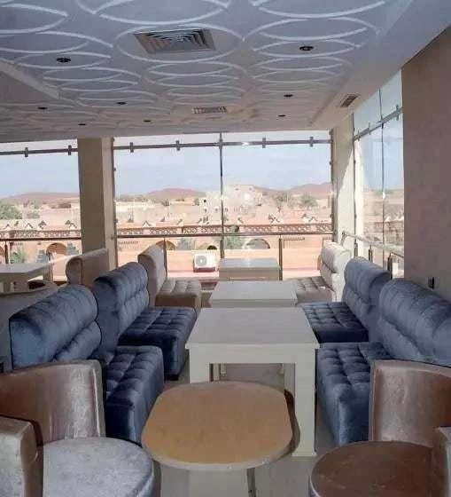Residence Touristique Merzouga lounge area with plush blue velvet sofas, beige armchairs, white tables, and large windows overlooking desert landscape