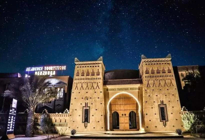 Residence Touristique Merzouga with traditional Moroccan architecture, beige stone walls, decorative patterns, and illuminated entrance at night under a starry sky.