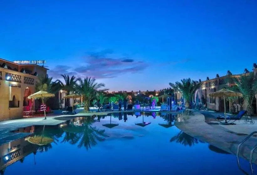 Residence Touristique Merzouga with illuminated outdoor pool, palm trees, straw umbrellas, and desert-style buildings at dusk