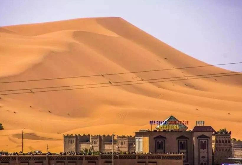 Residence Touristique Merzouga building with traditional architecture in front of large orange sand dunes under a clear sky