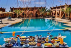 Residence Touristique Merzouga featuring a large blue swimming pool surrounded by terracotta buildings and a table with assorted breakfast foods and drinks