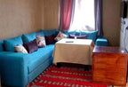 Residence Touristique Merzouga living room with turquoise upholstered corner sofa adorned with floral and solid cushions, beige tablecloth-covered wooden table, and red patterned carpet