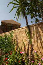 Riad Africa Marrakech with a woven bamboo fence, tall green palm trees, and red and pink flowering plants under a clear blue sky