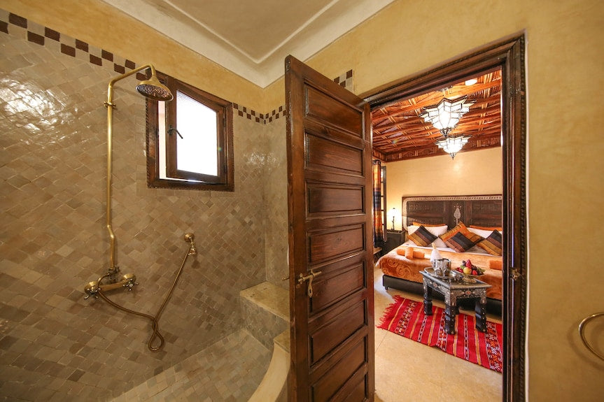 Riad Africa Marrakech bathroom with beige tiled walls, brass shower fixtures, wooden door opening to bedroom with decorative wood ceiling and colorful cushions