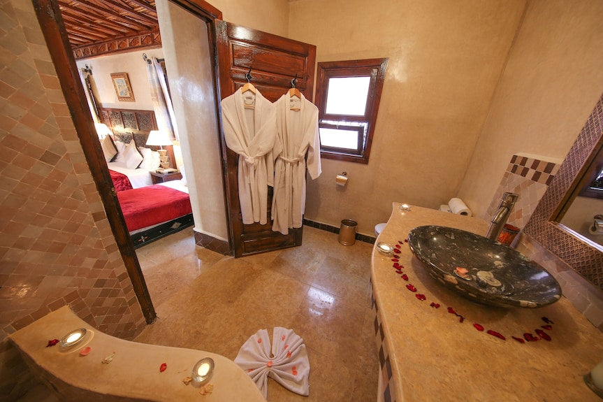 Riad Africa Marrakech bathroom with beige tiled walls, two white bathrobes hanging on wooden door, black stone sink, and rose petal decoration