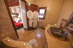 Riad Africa Marrakech bathroom with beige tiled walls, two white bathrobes hanging on wooden door, black stone sink, and rose petal decoration