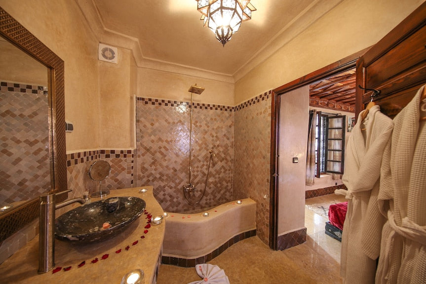 Riad Africa Marrakech bathroom with beige and brown tiled walls, stone sink, curved countertop, and hanging white robes
