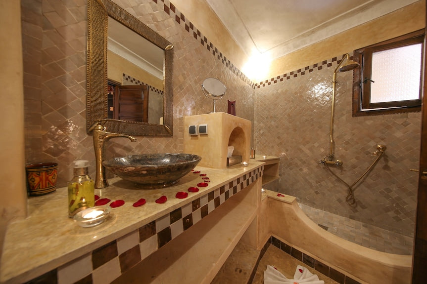 Riad Africa Marrakech bathroom with beige tiled walls, mosaic countertop, metallic sink, and brass fixtures, decorated with rose petals