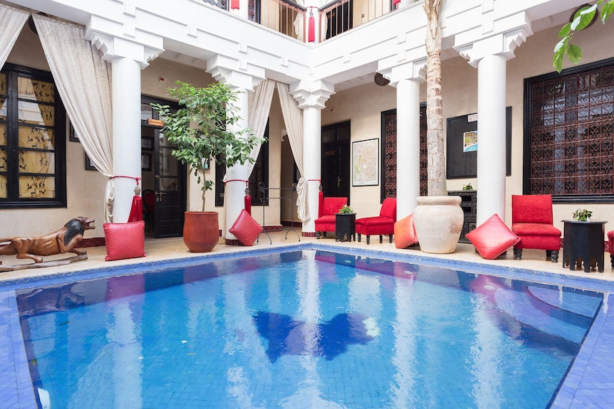 Riad Africa Marrakech courtyard with blue tiled swimming pool, white columns, beige walls, red cushioned chairs, and decorative plants